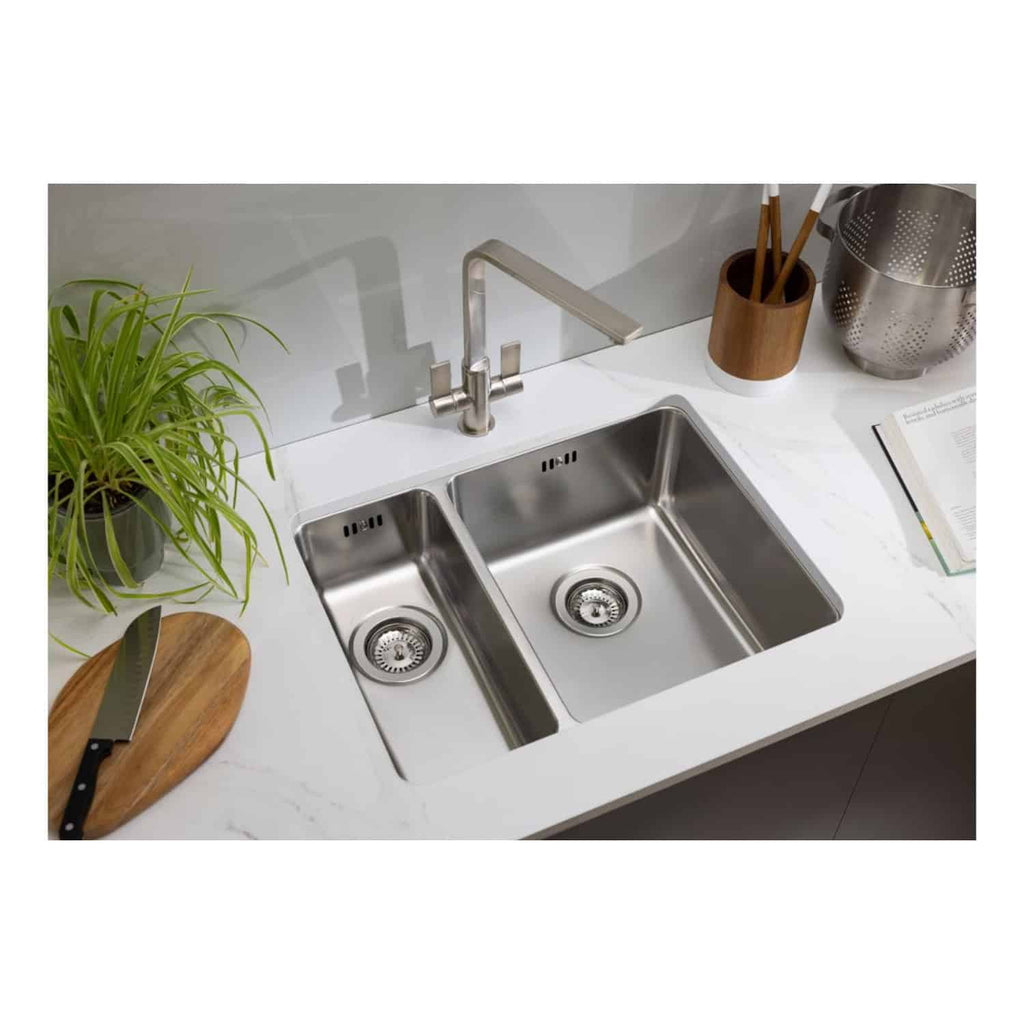 Stainless steel double kitchen sink with modern design, chrome faucet, and white marble countertop ideal for contemporary bathrooms and kitchens. Includes green plant, wooden cutting board, and kitchen accessories.