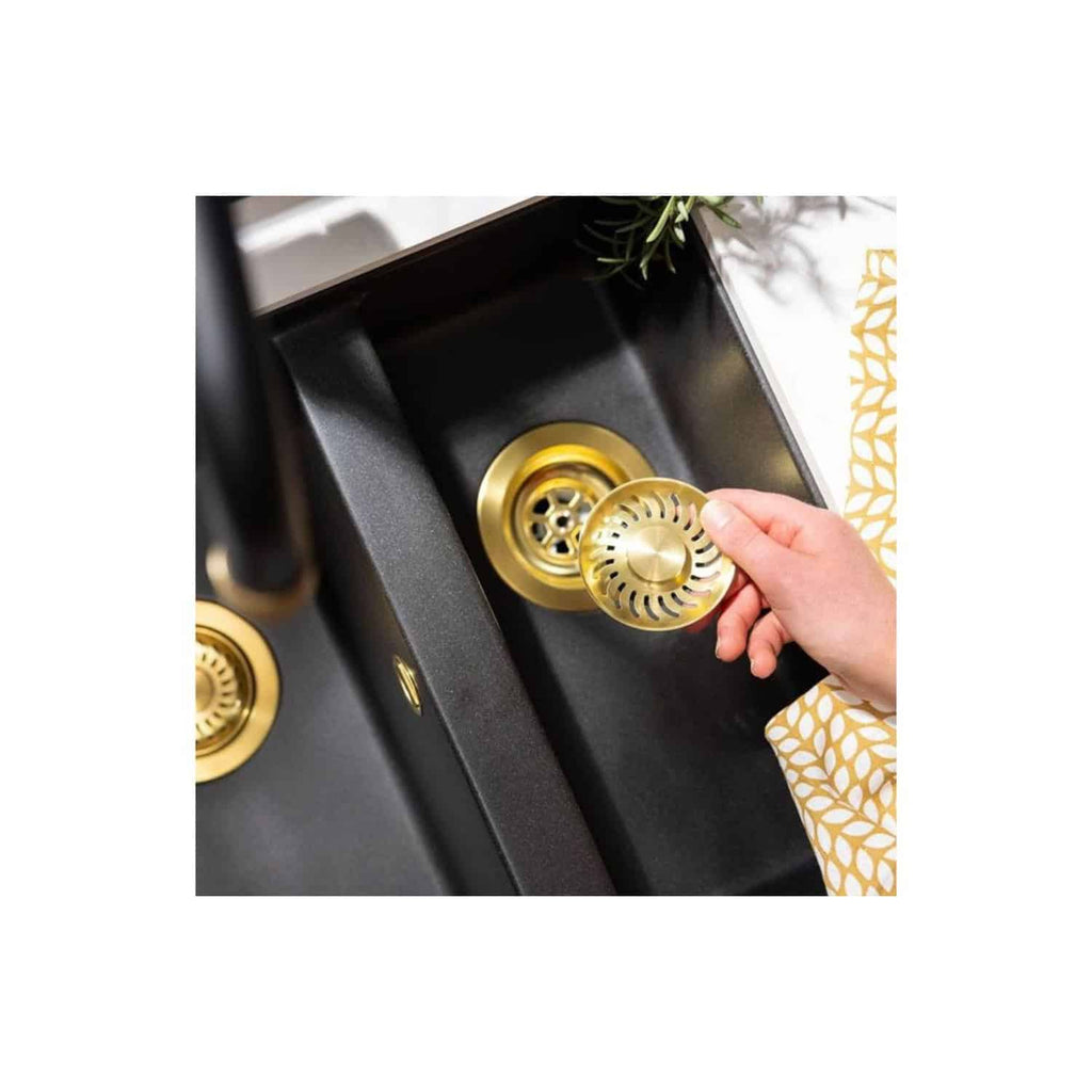 Polished gold sink strainer being installed in a modern black kitchen sink with gold accents and a decorative plant in the background.
