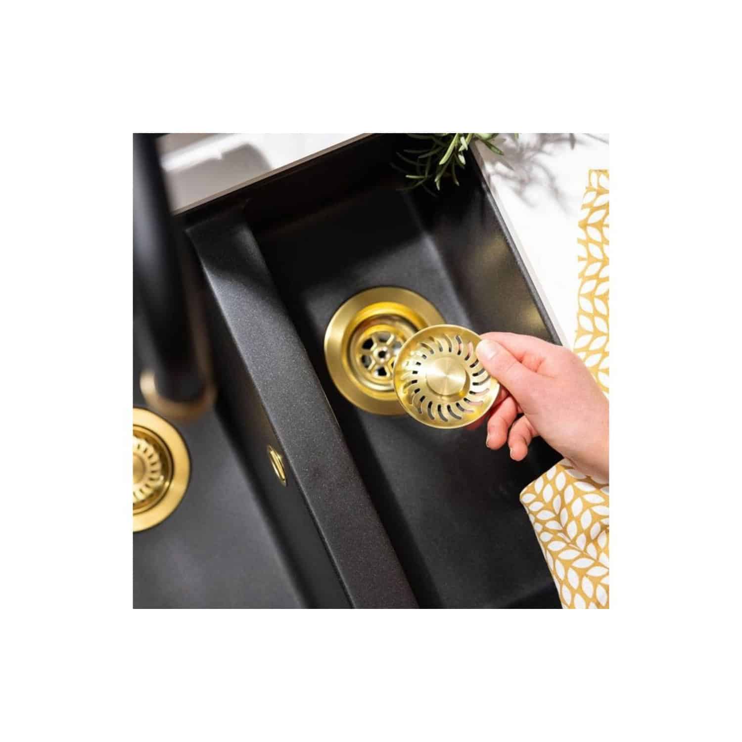 Polished gold sink strainer being installed in a modern black kitchen sink with gold accents and a decorative plant in the background.