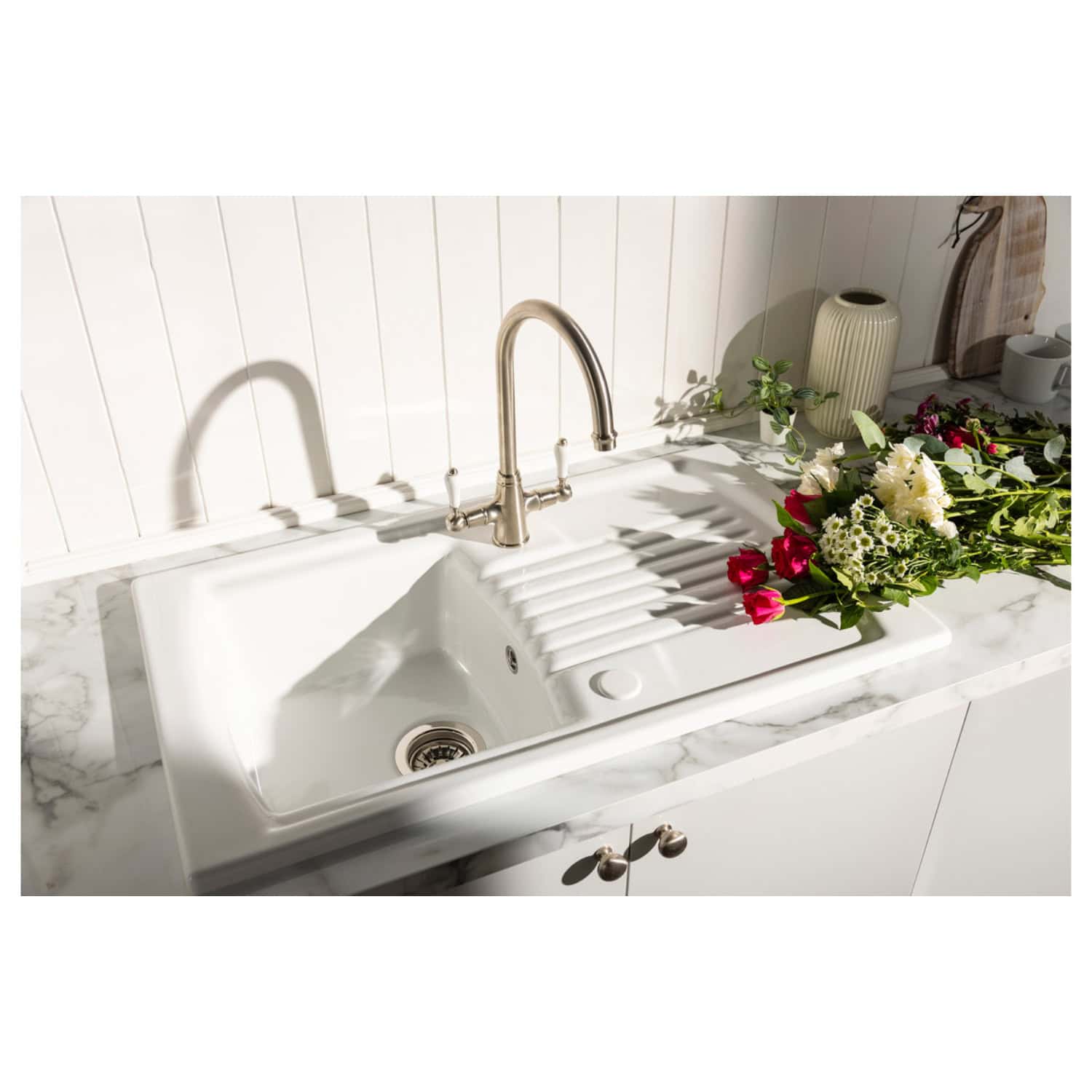 Shiny stainless steel kitchen tap above white farmhouse ceramic sink with draining board, set on white marble countertop, with fresh flowers and decorative vases in a modern bathroom or kitchen setting.
