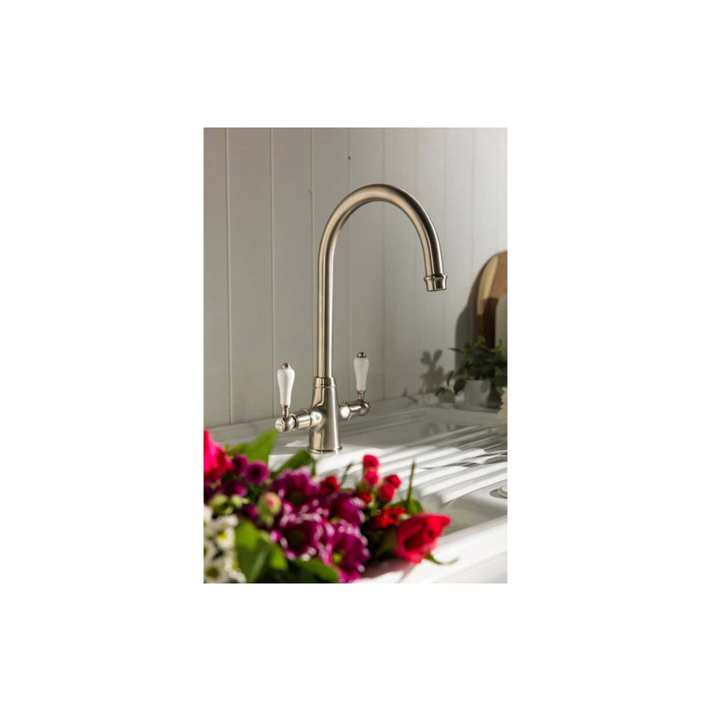 Polished stainless steel kitchen sink faucet with dual white ceramic handles, modern design, installed in a light grey wooden wall kitchen or utility room setting.