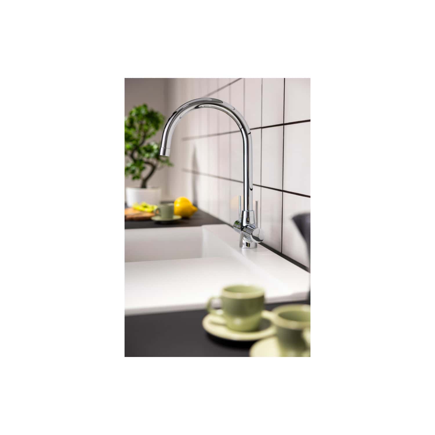 A sleek, modern chrome kitchen faucet installed over a white sink in a contemporary kitchen setting. The background features beige tiled walls, a potted plant, and a yellow lemon along with cups and saucers for an inviting look.