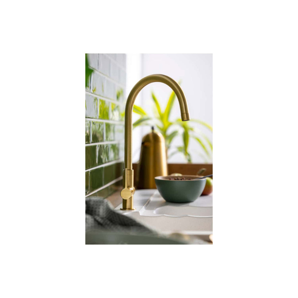 Shiny gold kitchen tap with a modern high-arch design against a green tiled backsplash, surrounded by a bowl of cereal, fruit, and houseplants, creating a stylish and inviting kitchen space.