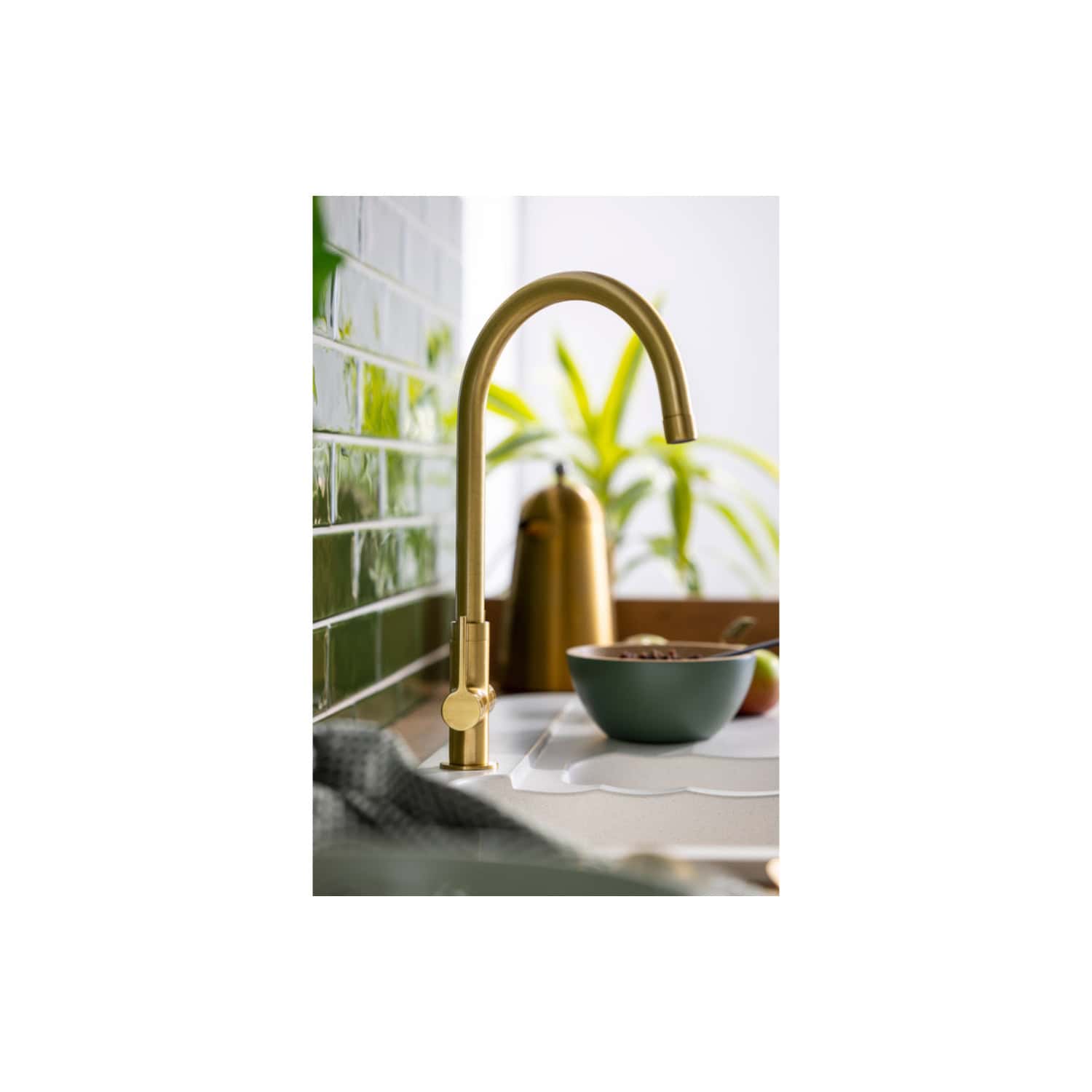 Shiny gold kitchen tap with a modern high-arch design against a green tiled backsplash, surrounded by a bowl of cereal, fruit, and houseplants, creating a stylish and inviting kitchen space.