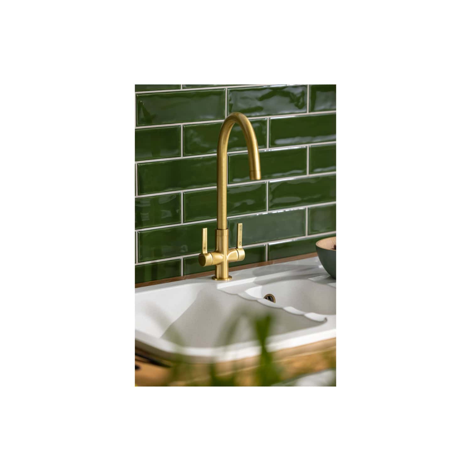 Brass kitchen tap with a modern design installed above a white sink with scalloped edges, set against a dark green subway tile backsplash, creating a stylish and contemporary kitchen look.