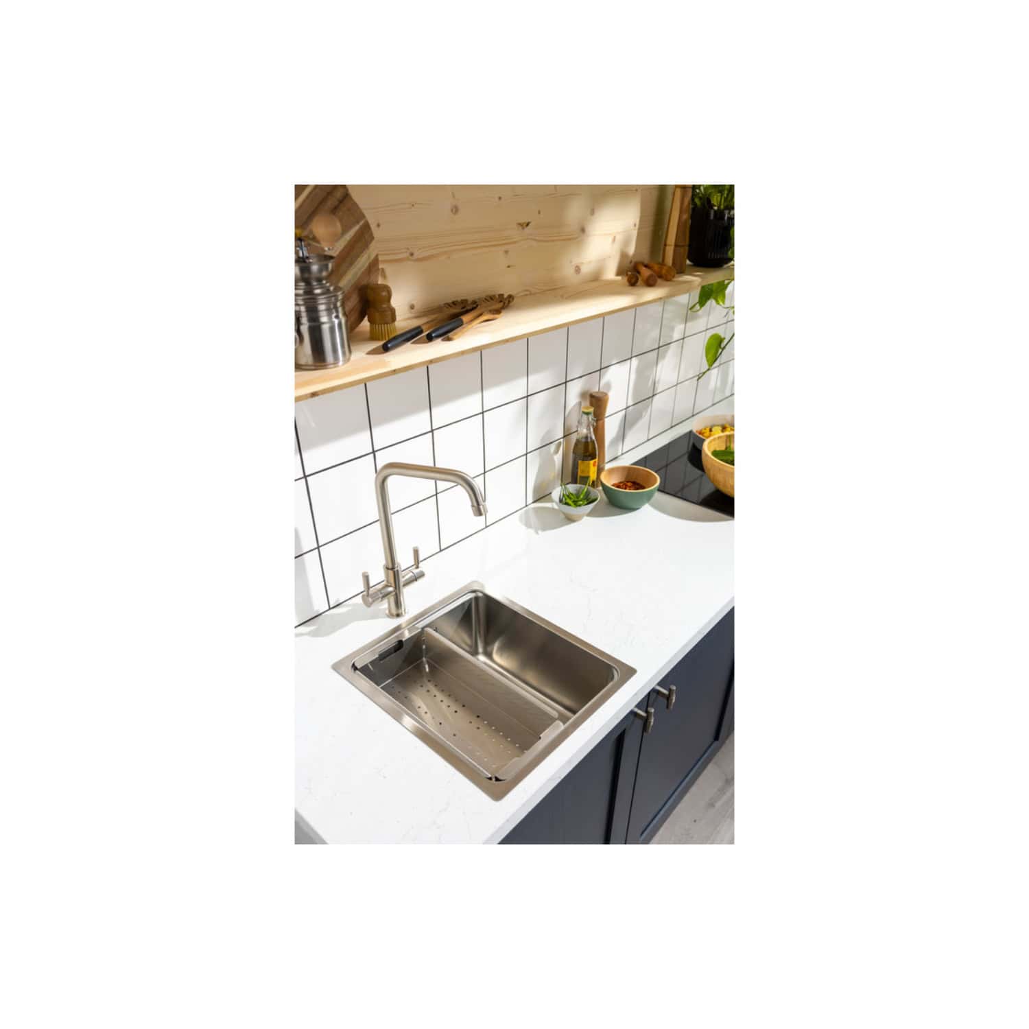 Shiny stainless steel kitchen sink with modern chrome faucet, set in a sleek white countertop and dark blue base cabinets, with a backdrop of white tiled wall and natural wooden shelf, ideal for contemporary bathroom or kitchen design.