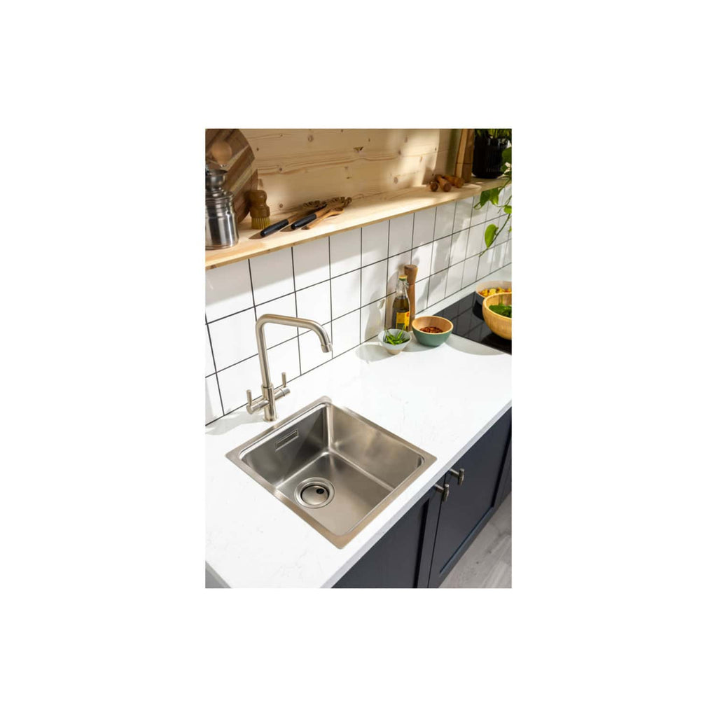 Shiny stainless steel kitchen sink with modern tap mounted on a white countertop, set against a white tiled backsplash and wooden open shelving with kitchen essentials and decorative items.