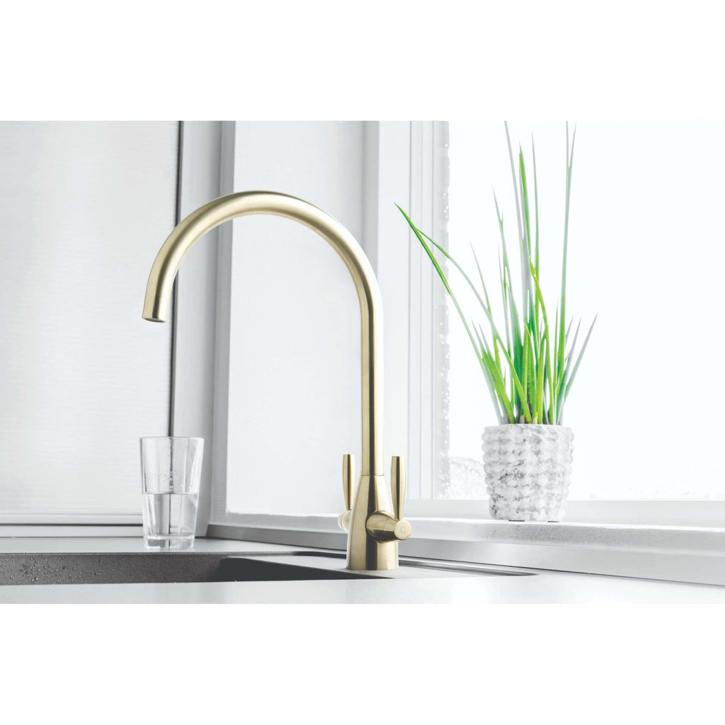 Sleek gold kitchen tap with a modern curved design, positioned over a sink next to a clear glass of water and a decorative white textured vase with green grasses on a bright windowsill.