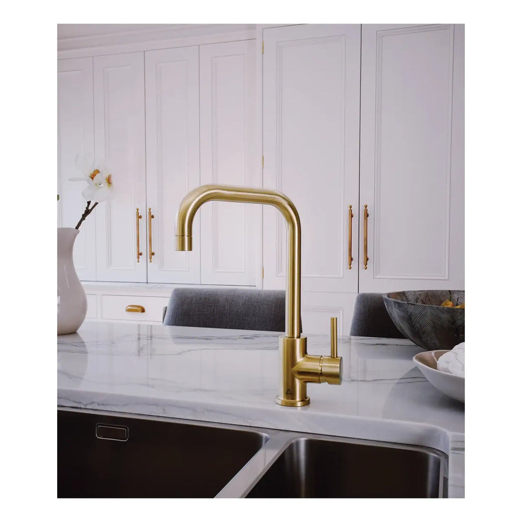 Shiny gold kitchen tap with a modern curved design, installed on a white marble countertop in a sleek, contemporary kitchen setting.