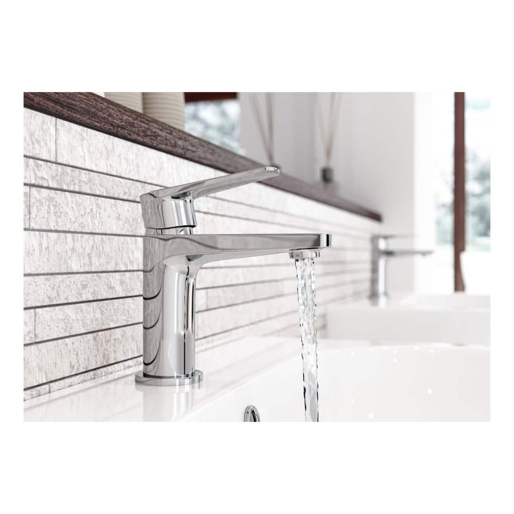 Brushed chrome bathroom mixer tap with water running over a modern white sink, reflected in natural light. Stylish, contemporary bathroom fixture surrounded by textured white tiles.