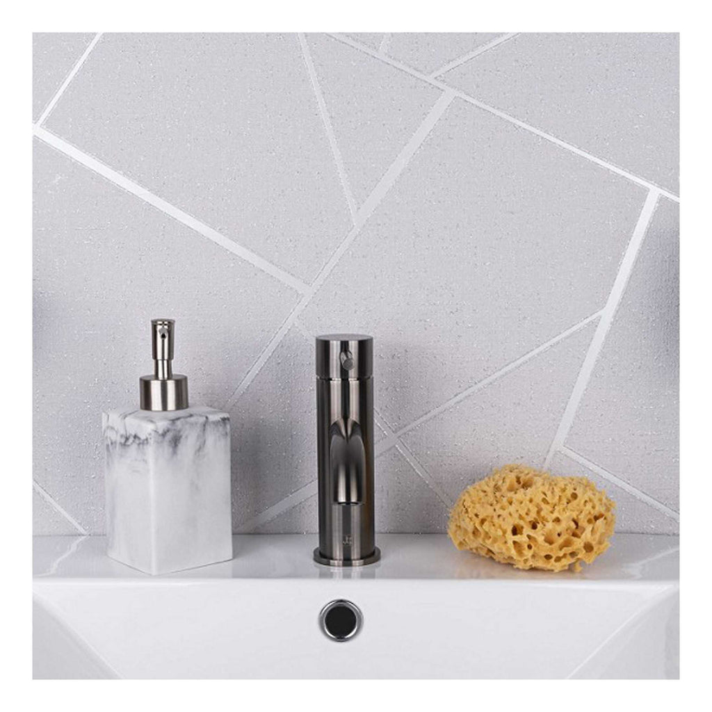 Polished chrome bathroom tap with soap dispenser and cleaning sponge on a white sink against textured, geometric-patterned grey tile wall, modern and minimalist bathroom design.