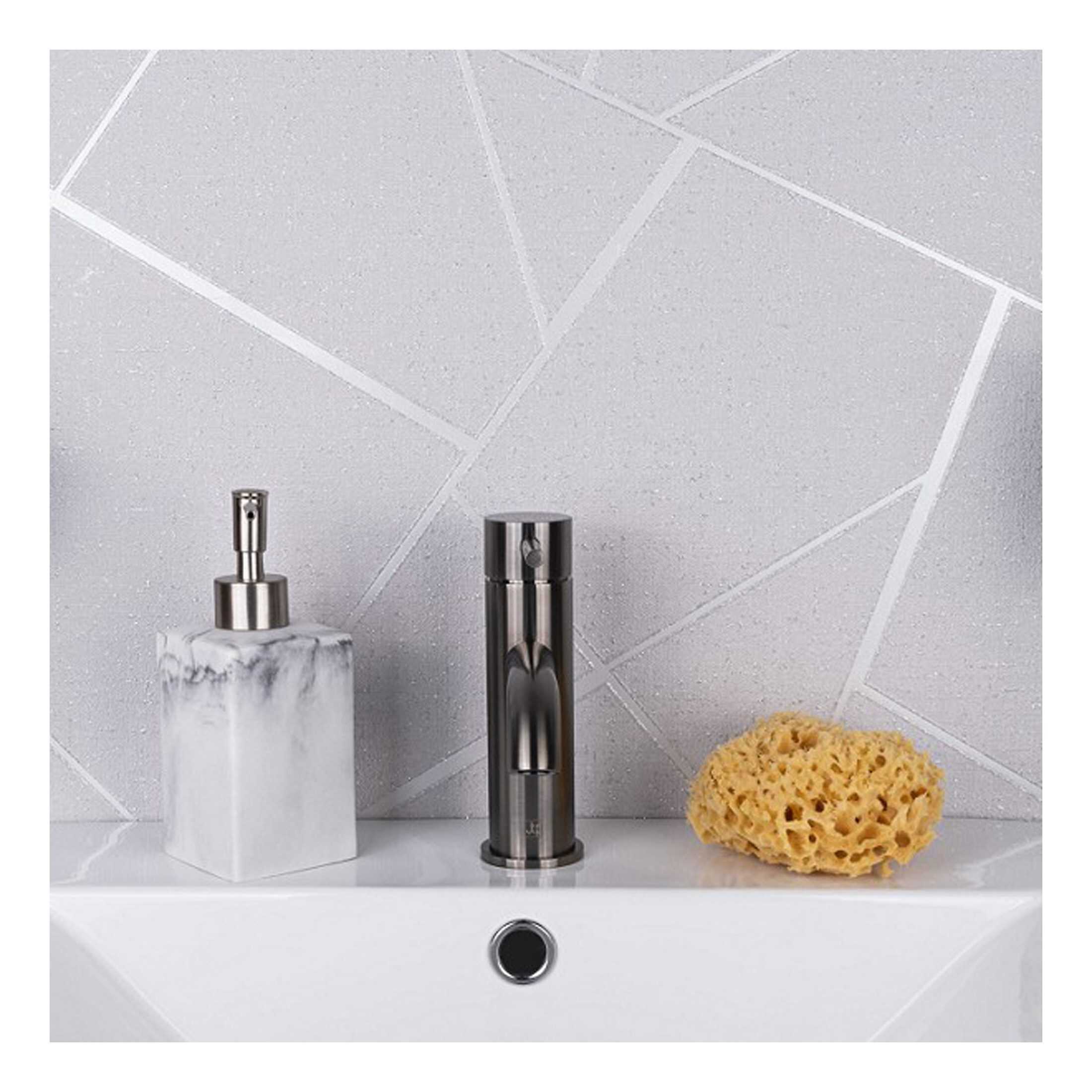 Polished chrome bathroom tap with soap dispenser and cleaning sponge on a white sink against textured, geometric-patterned grey tile wall, modern and minimalist bathroom design.
