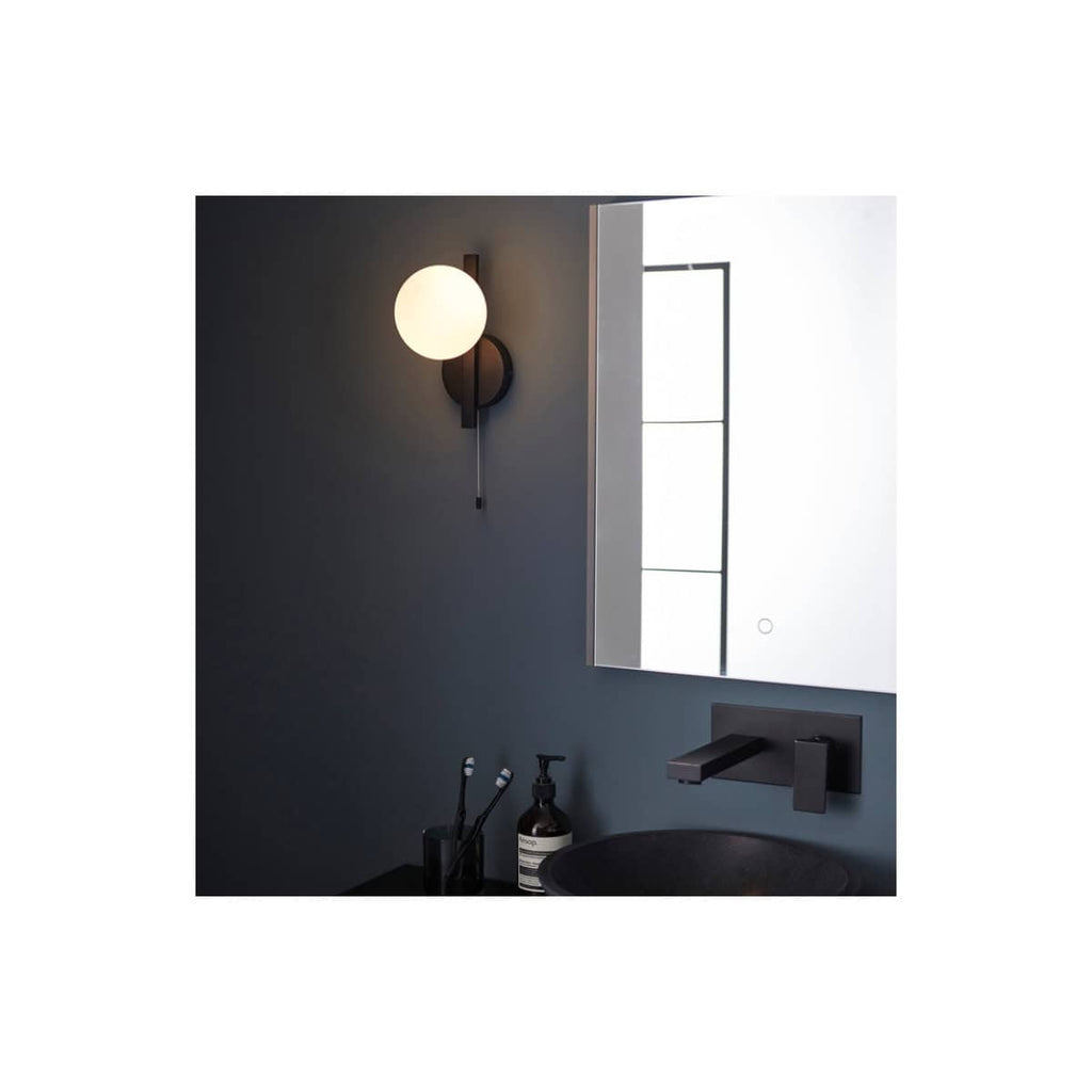 Modern black wall-mounted bathroom light fixture with a spherical white globe, installed beside a sleek mirror and minimalist black faucet, creating a contemporary bathroom ambiance.
