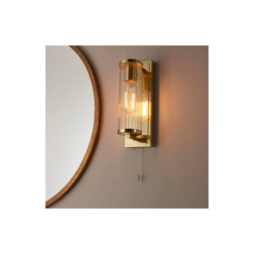 Elevated modern bathroom wall light fixture with a cylindrical glass shade and sleek brass accents, providing ambient illumination in contemporary bathroom designs.