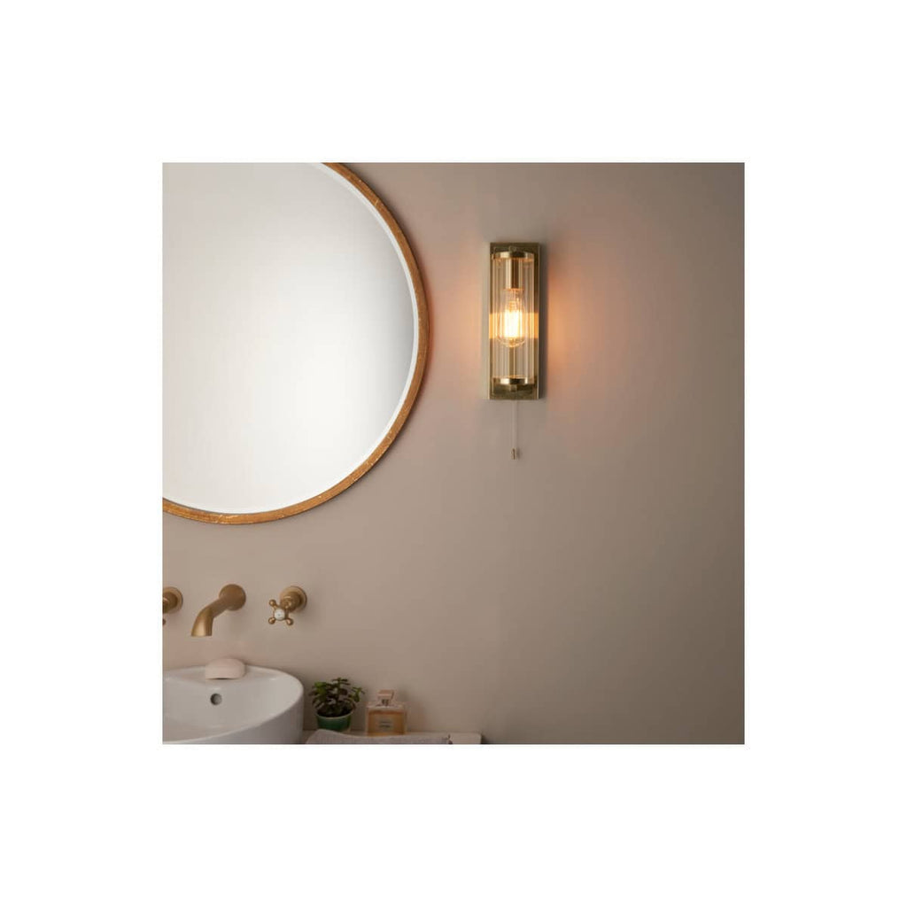 Elegant bathroom wall light fixture with brass accents illuminating a modern bathroom with a round mirror and minimalist decor. Highlights stylish lighting for contemporary bathroom design.