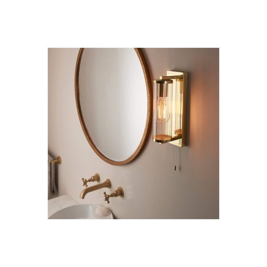 Sleek modern bathroom wall sconce with amber glass shade, mounted beside a large oval wooden-framed mirror, featuring traditional gold-tone fixtures and soft lighting for elegant bathroom decor.