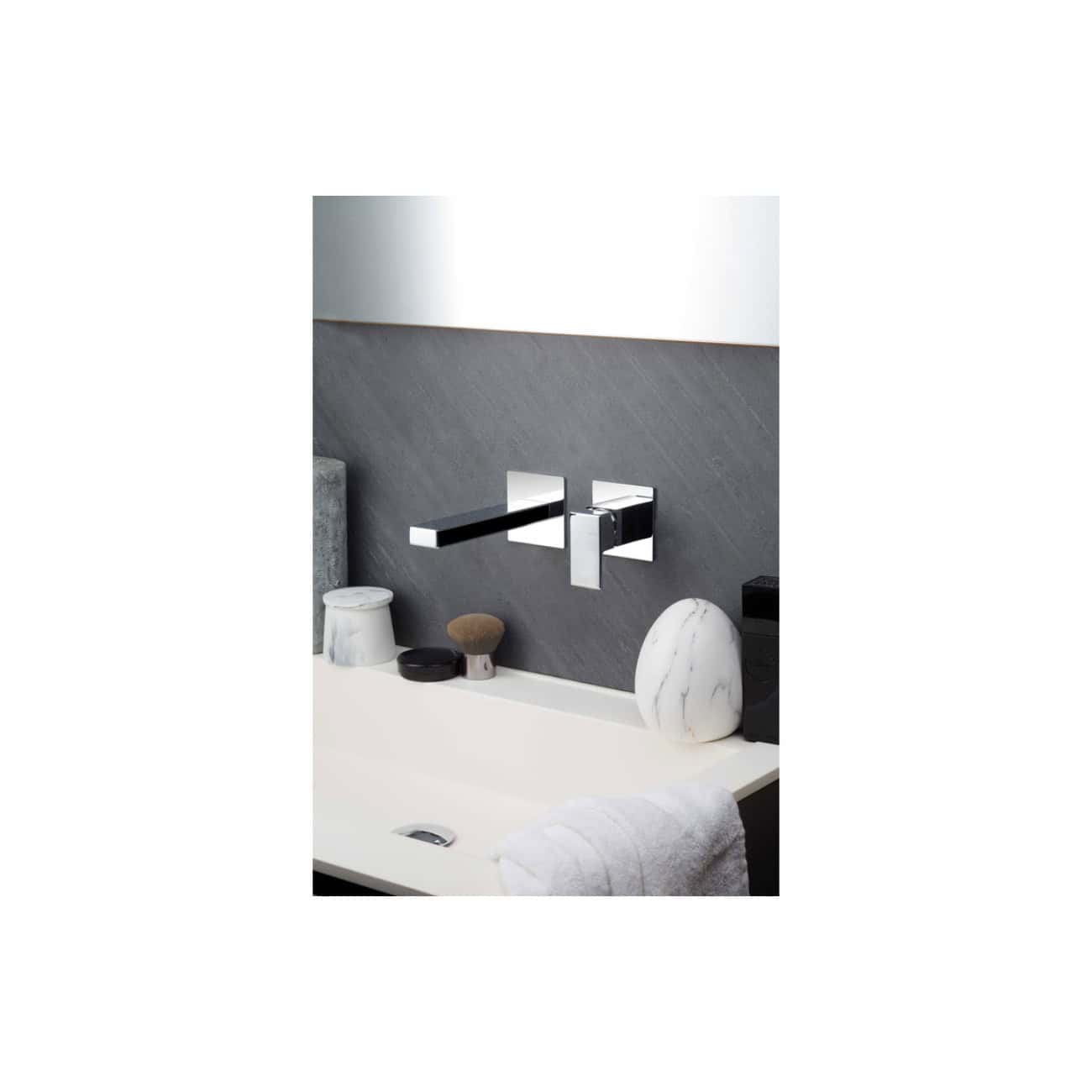 Sleek modern chrome bathroom wall-mounted faucet with a minimalist design, installed over a stylish grey stone backsplash and white sink; complemented by decorative stones and accessories.