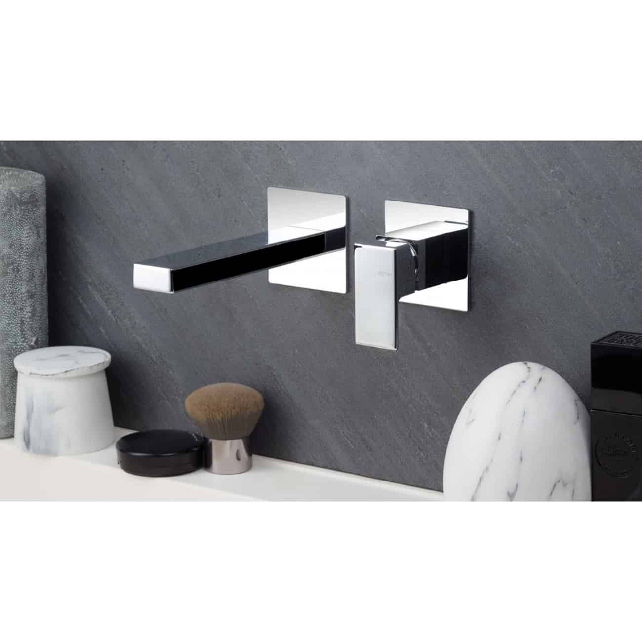 Polished chrome wall-mounted bathroom tap with modern geometric design on grey textured wall.