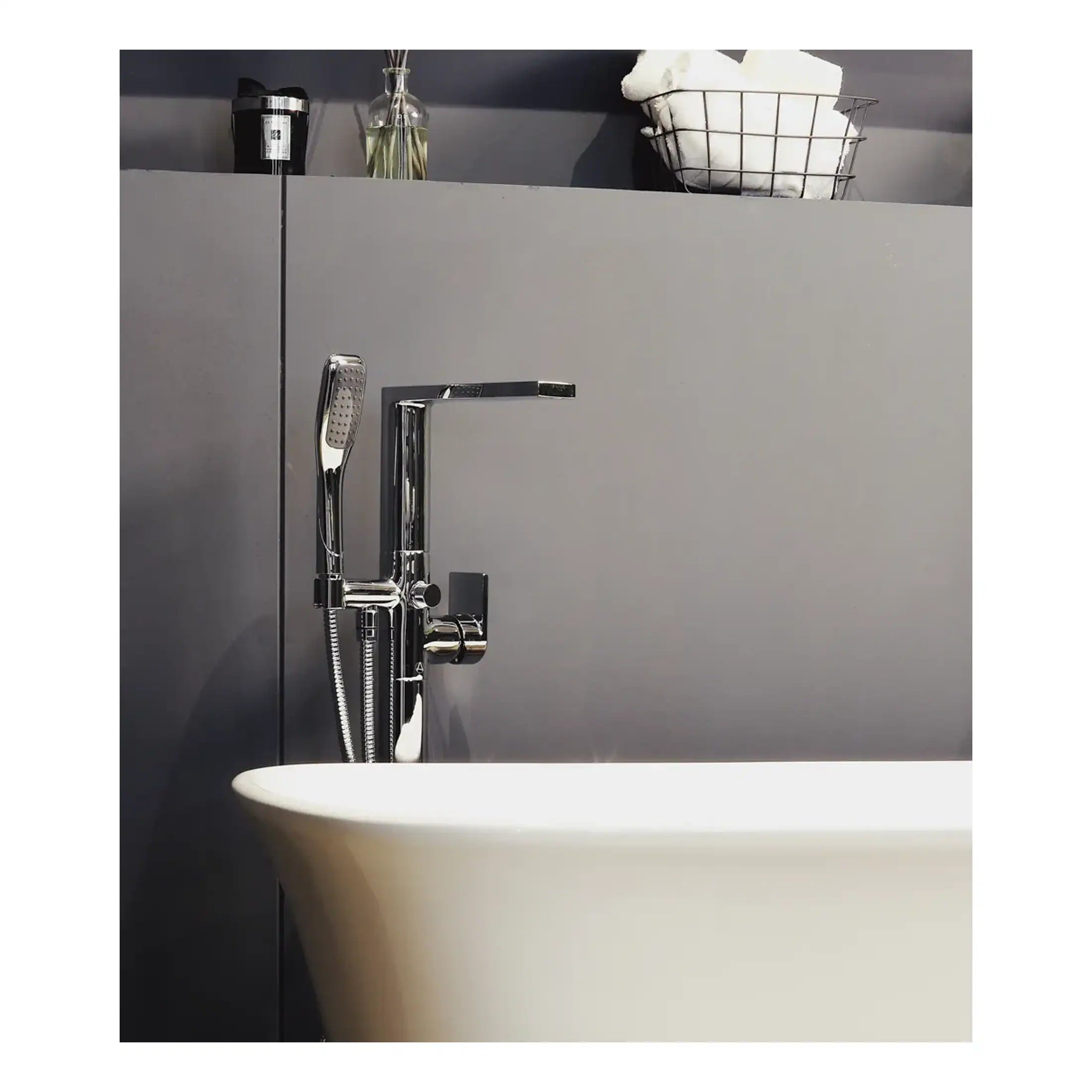 Sleek modern bathroom tap and handheld shower attached to a white bathtub, with minimalist grey wall and shelving featuring towels and toiletries, representing contemporary bathroom design.