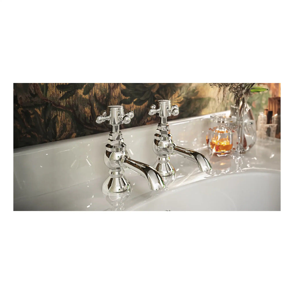Elegant Victorian-style chrome bathroom taps on a white ceramic sink with decorative toiletries and floral arrangements in the background, showcasing luxury bathroom fixtures and fittings.