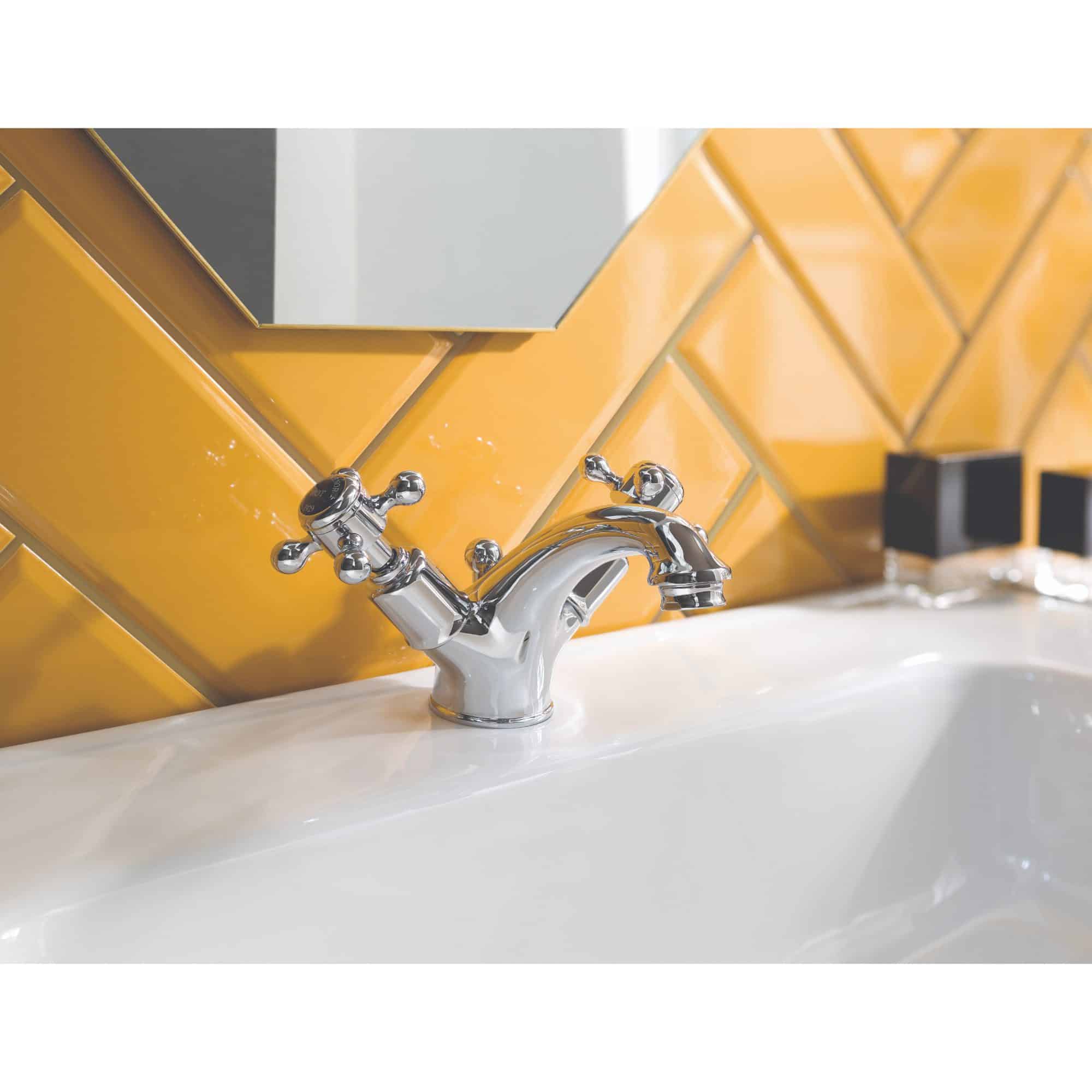 Shiny chrome bathroom tap with dual handles mounted on a white sink, set against a modern yellow tiled wall, highlighting contemporary bathroom fixtures and stylish design.