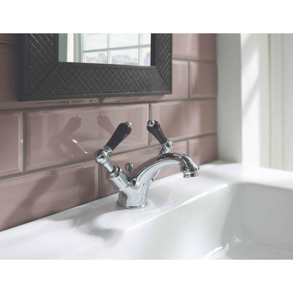 Elegant chrome bathroom faucet with dual handles on a white sink, set against pink tiled wall and a black-framed mirror, showcasing stylish and functional bathroom design.