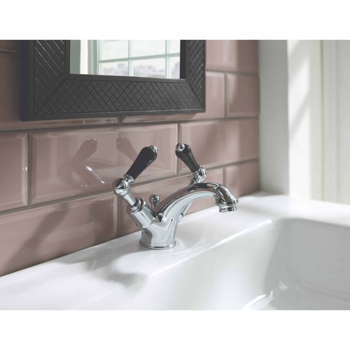 Elegant chrome bathroom faucet with dual handles on a white sink, set against pink tiled wall and a black-framed mirror, showcasing stylish and functional bathroom design.
