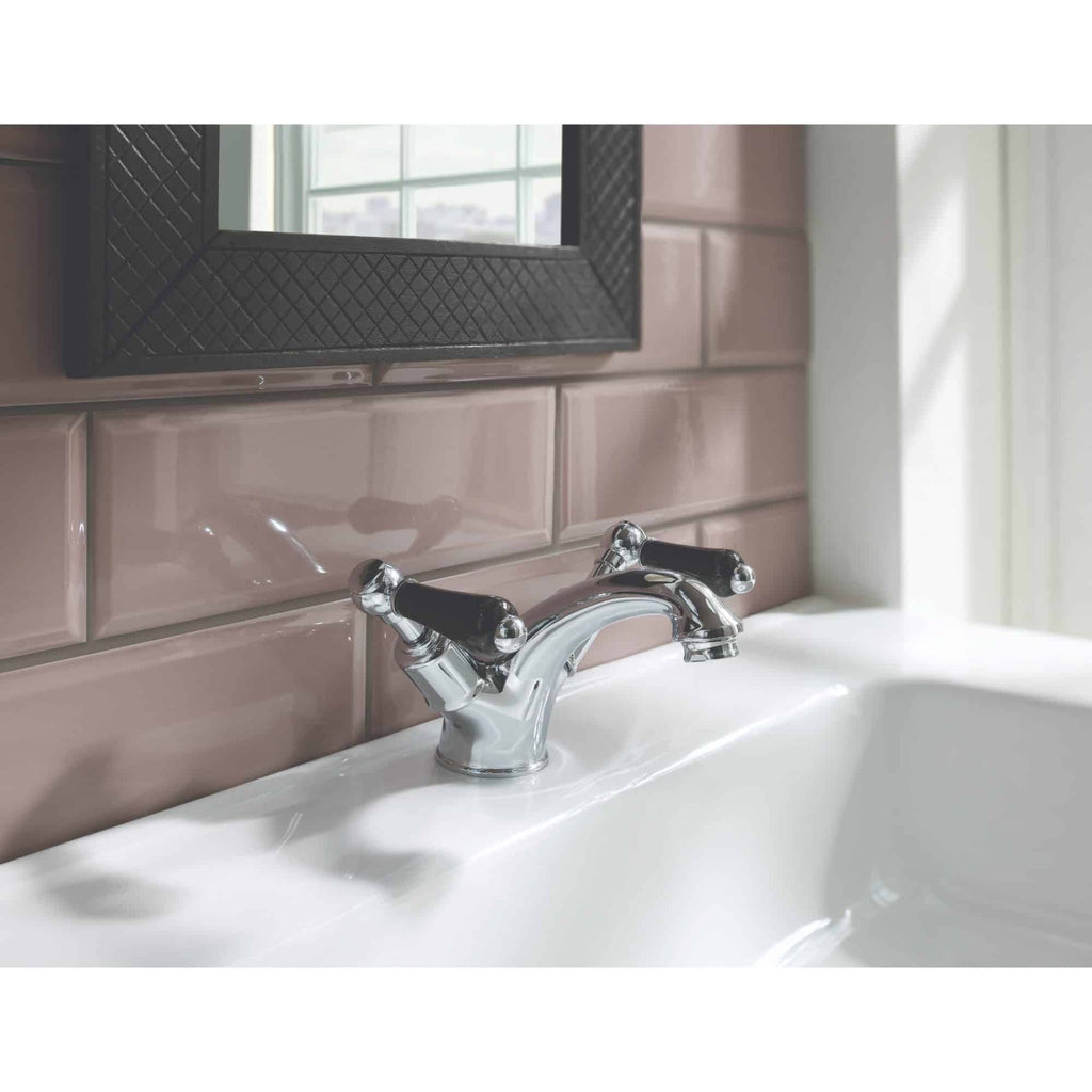 Shiny chrome bathroom sink faucet with black handles mounted on a white ceramic sink, pink glossy tiled wall, and a black textured mirror frame reflecting natural light, showcasing elegant bathroom fixtures.