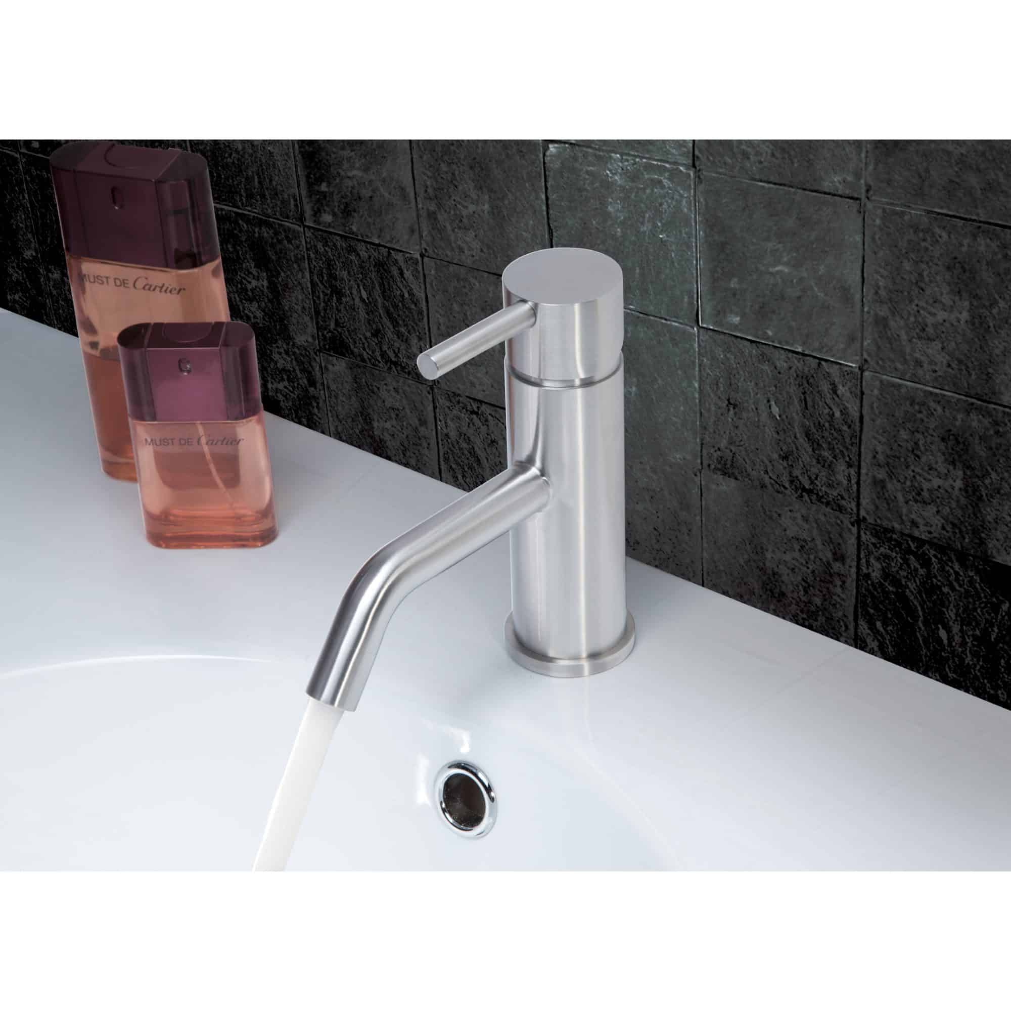 Sleek modern stainless steel bathroom sink faucet with water flowing, set against black tiled wall with beauty products nearby.