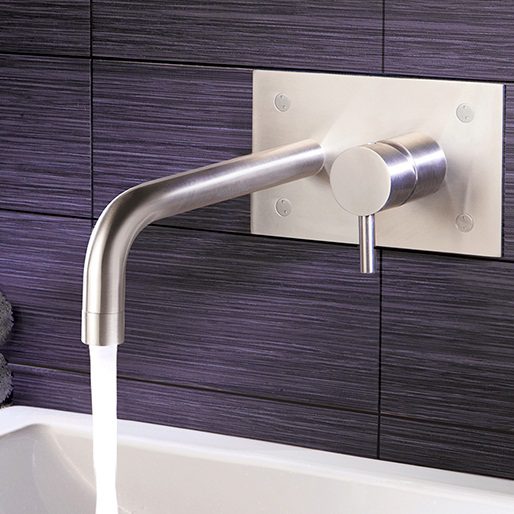 Sleek modern stainless steel bathroom tap with water running, mounted on dark tiled wall behind a white bathroom basin.