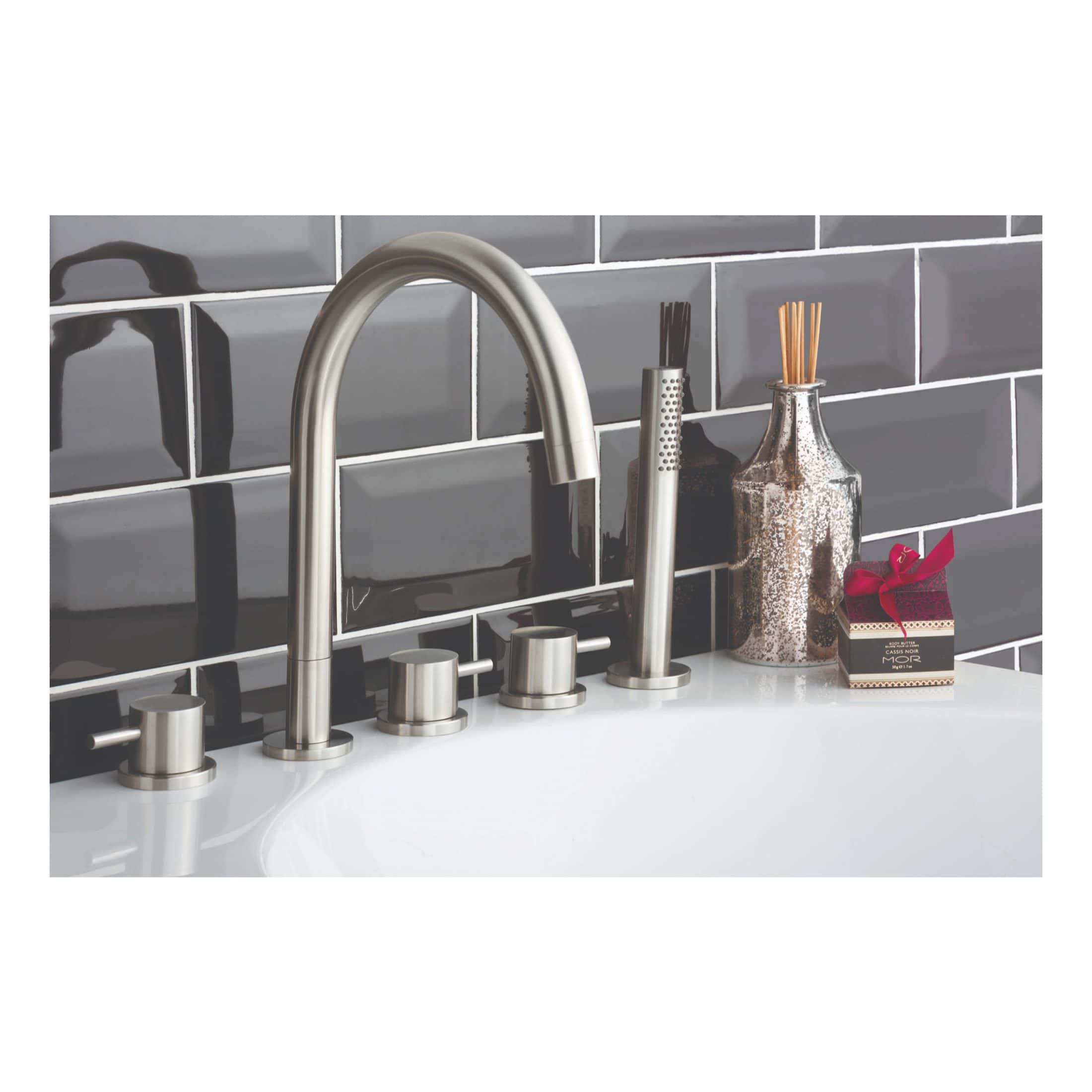 Polished stainless steel kitchen tap with modern design, alongside matching soap dispenser, black diffuser, decorative vase with reed sticks, and a gift box, set against dark grey subway tiles for a stylish bathroom look.