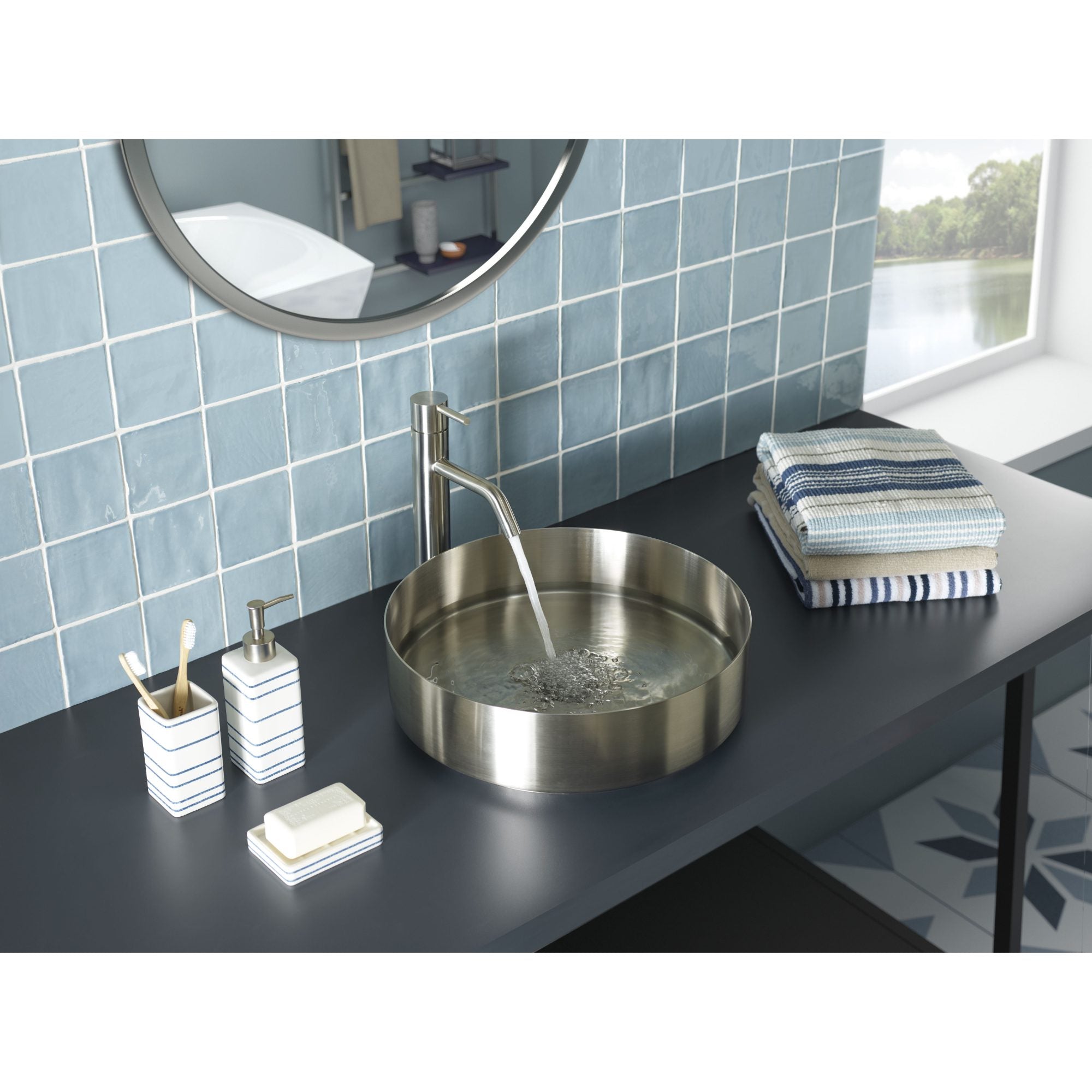 Shiny stainless steel bathroom sink with modern faucet on a black countertop, blue tiled wall, neatly folded towels, and soap dispenser, set in a contemporary bathroom with large window overlooking nature.