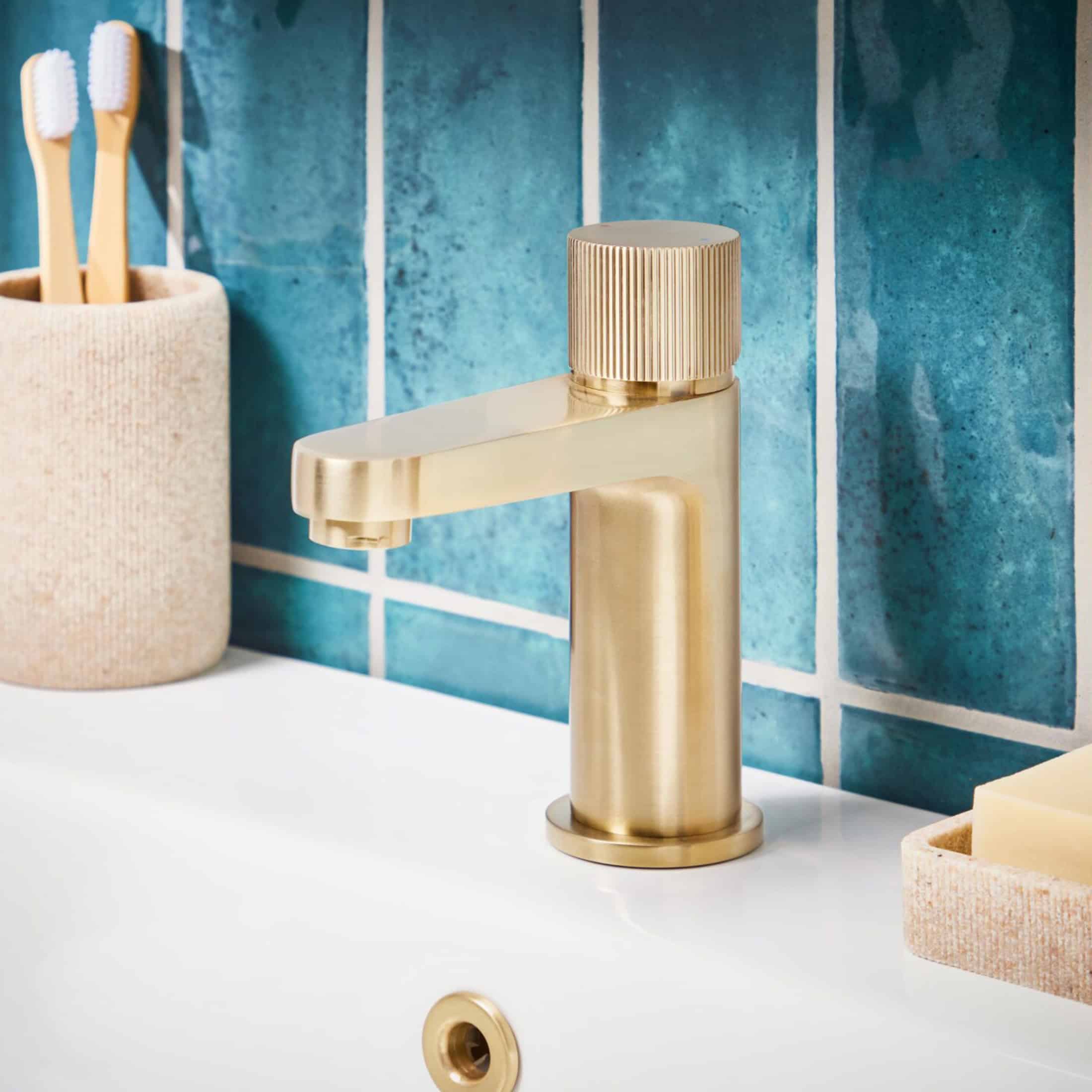 Sleek gold bathroom tap with modern design on a white sink, blue tiled wall background, and bathroom accessories, perfect for contemporary bathroom renovations or upgrades.