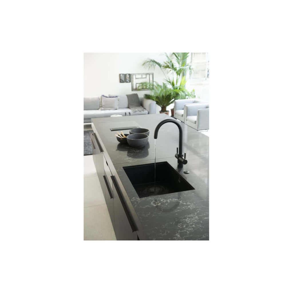 Sleek black kitchen tap with flowing water over a black marble kitchen island, modern open-plan living space with grey sofa, white chairs, and lush indoor plants in the background, stylish and contemporary kitchen design.