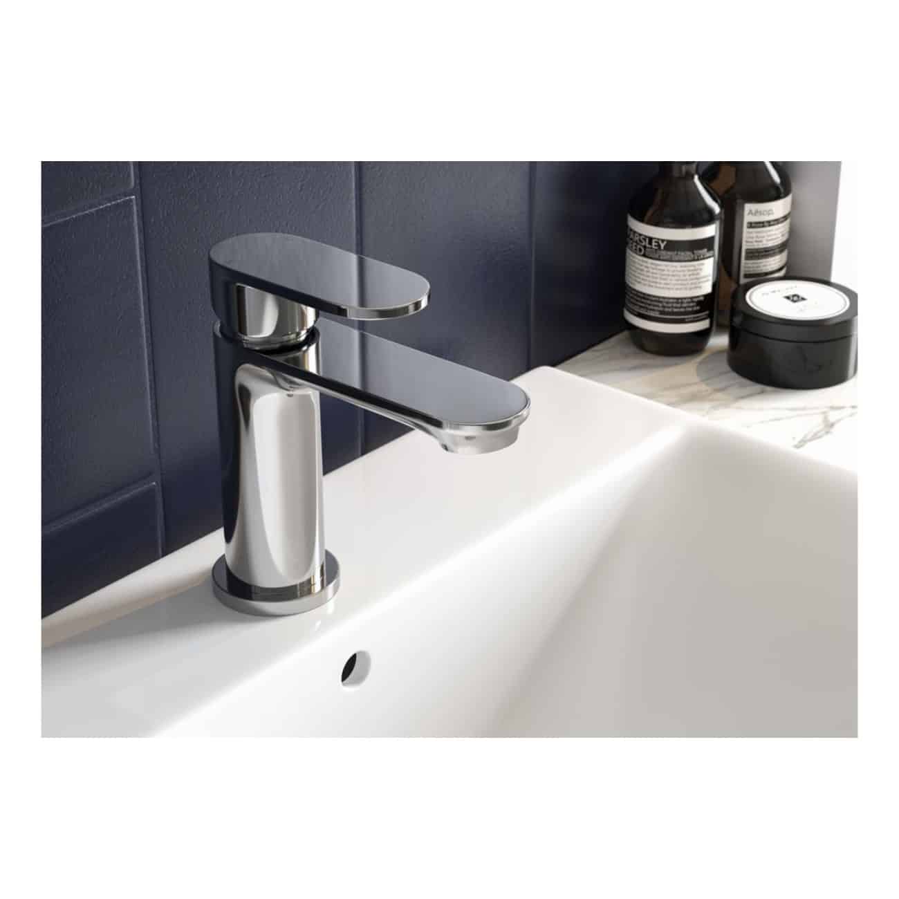 Sleek chrome bathroom tap on white ceramic sink with dark navy tiled wall background, modern bathroom fixtures, and personal care products on marble countertop.