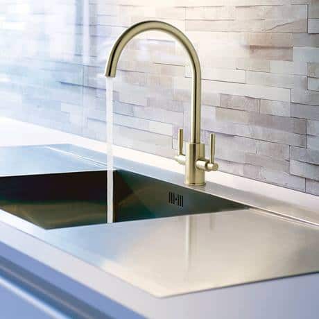 Sleek modern stainless steel kitchen sink with gold-toned bridge tap, set against a textured tiled wall, showcasing elegant bathroom and kitchen fixtures by Bubly Bathrooms.