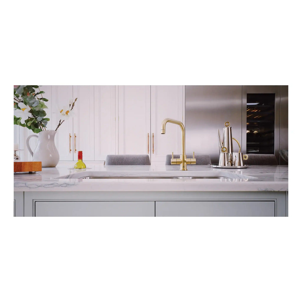 Shiny gold kitchen tap with modern design on a white marble countertop in a contemporary kitchen setting. Elegant accessories and a fresh floral arrangement add style to the space.