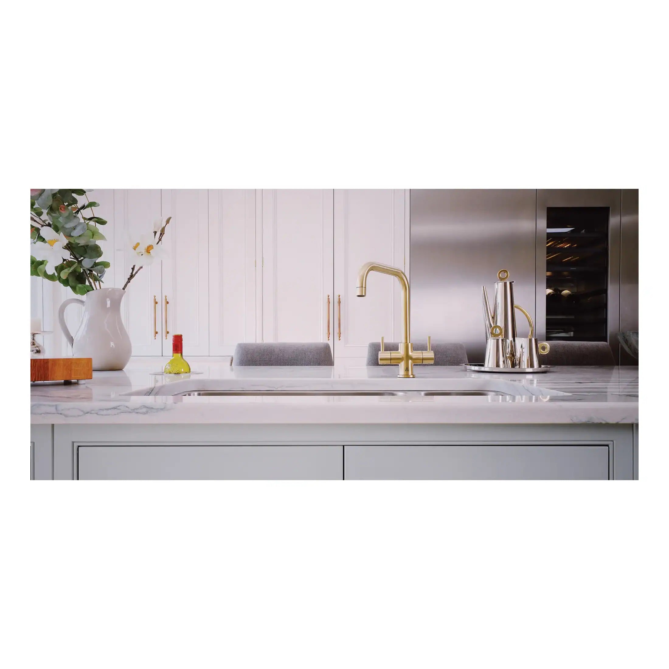 Shiny gold kitchen tap with modern design on a white marble countertop in a contemporary kitchen setting. Elegant accessories and a fresh floral arrangement add style to the space.