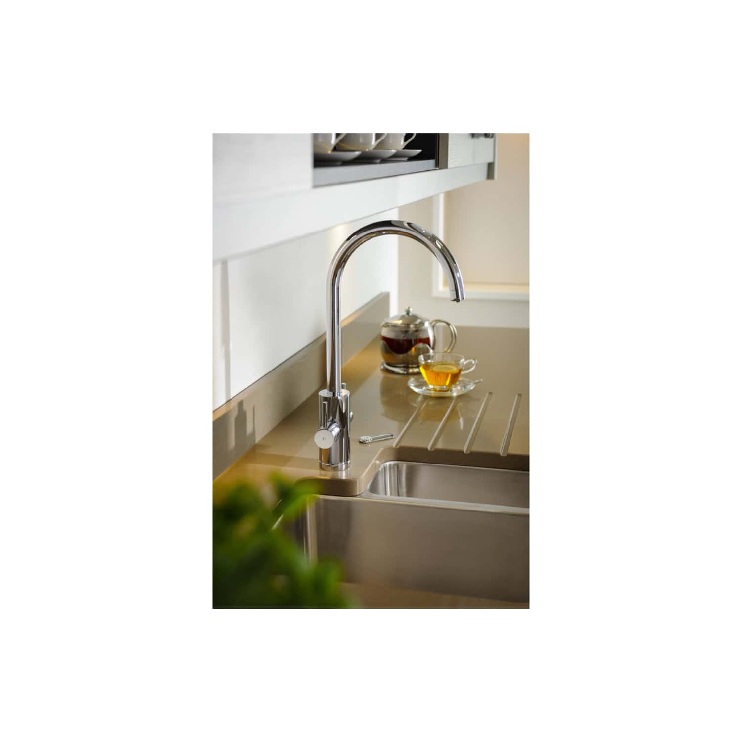 Shiny chrome kitchen faucet with a modern design, installed over a stainless steel sink, situated in a contemporary kitchen setup with a beige countertop and a teapot with tea and a cup of tea nearby.