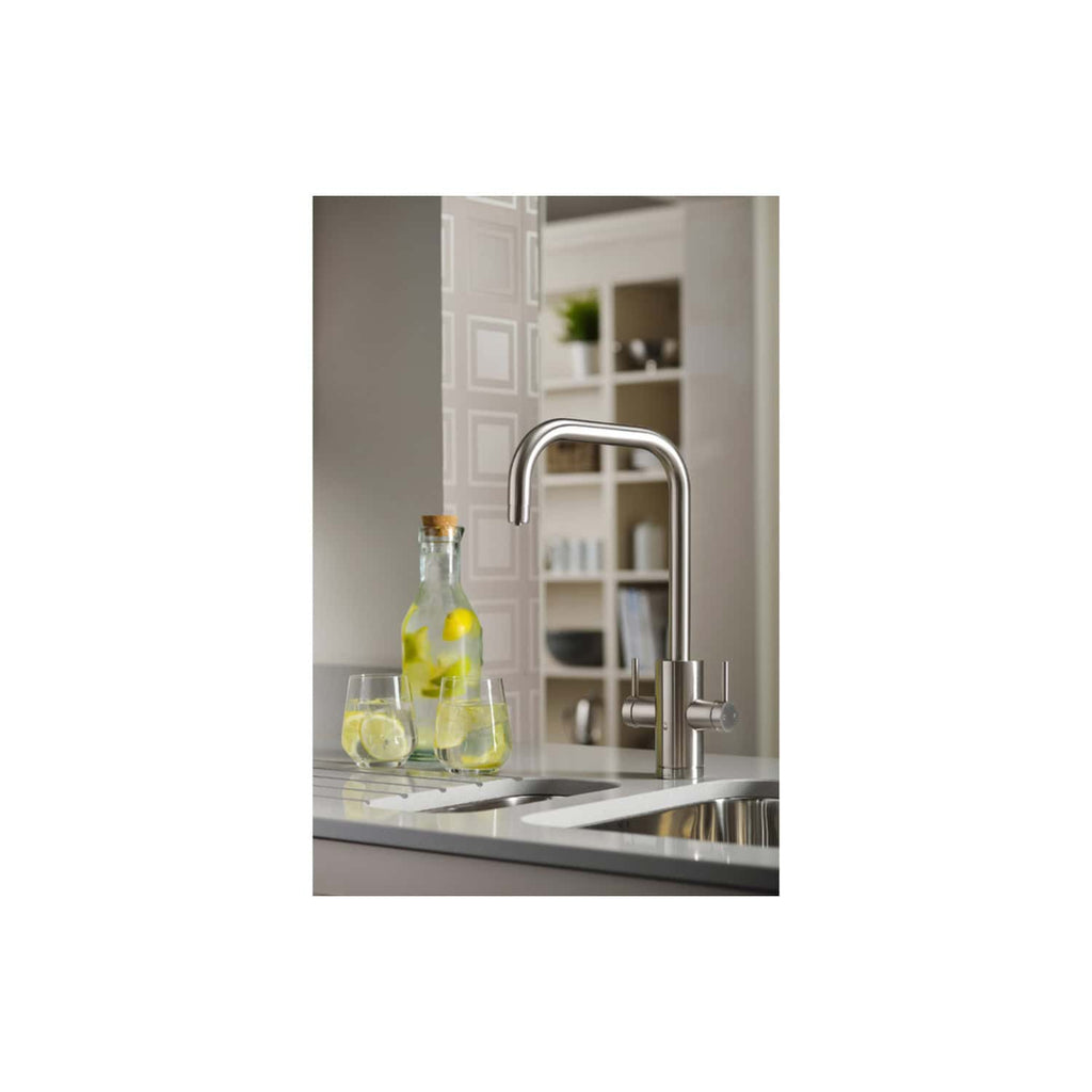 Futuristic stainless steel kitchen tap with curved spout in a modern kitchen setting. Bottles with lemon-infused water and glasses are placed on the white countertop under the tap. Shelves with decorative items are visible in the background.