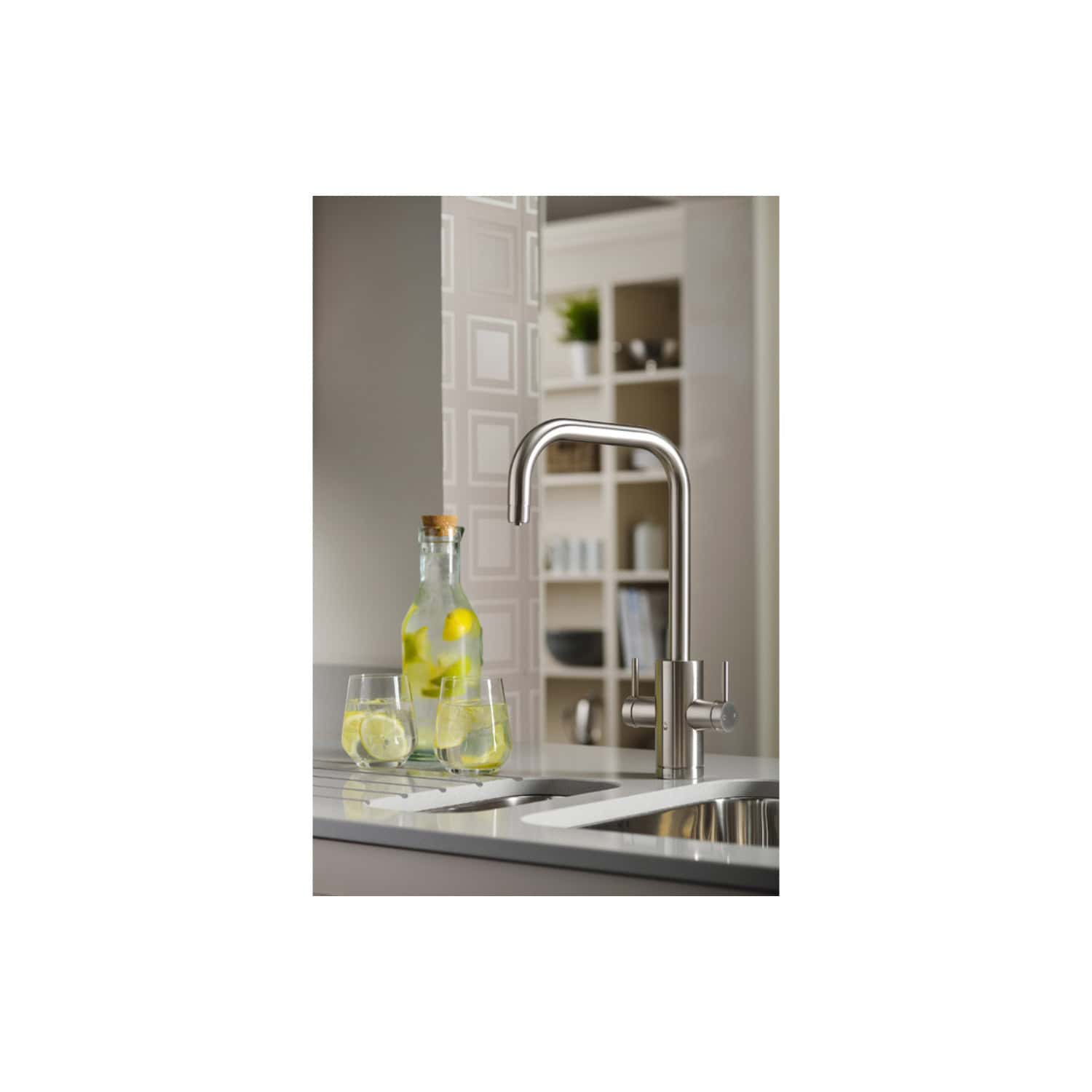 Futuristic stainless steel kitchen tap with curved spout in a modern kitchen setting. Bottles with lemon-infused water and glasses are placed on the white countertop under the tap. Shelves with decorative items are visible in the background.