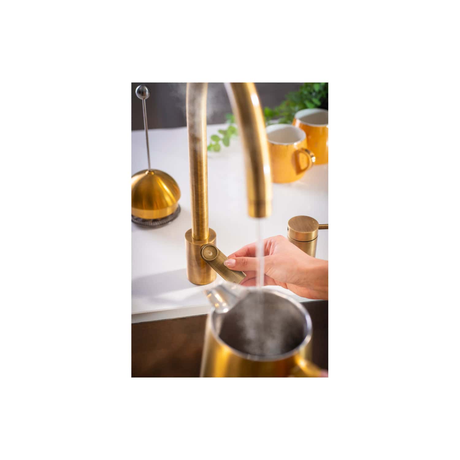 Polished gold kitchen tap with a modern lever design, in use with water flowing, featuring nearby cups and green plants, showcasing stylish bathroom fittings and fixtures.