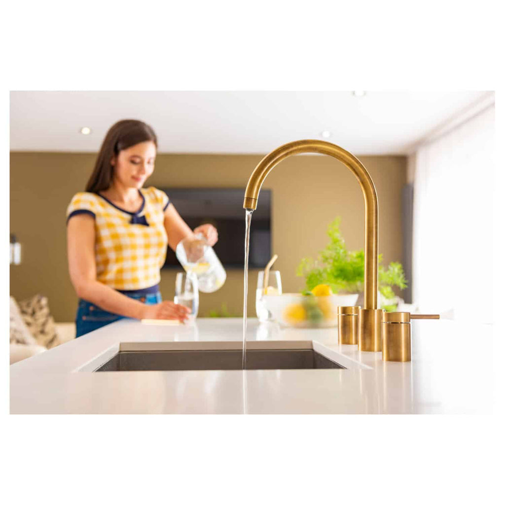 Shiny gold kitchen tap with running water in a modern kitchen setting, featuring a woman pouring water into a glass, stainless steel sink, and fresh green herbs on the counter.
