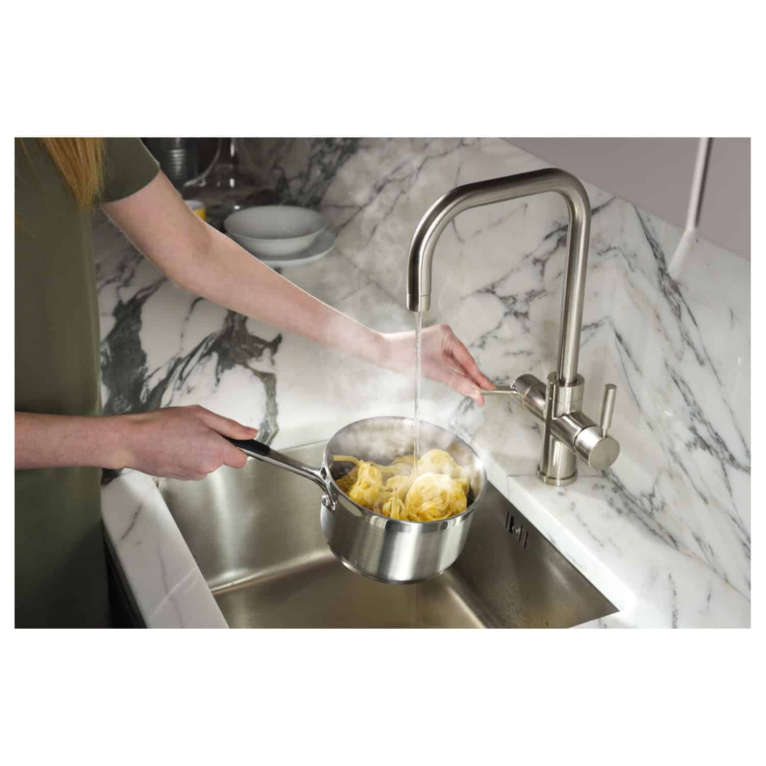 Feminine hand under modern kitchen faucet with running water, washing pasta in a stainless steel pot on a marbled countertop.