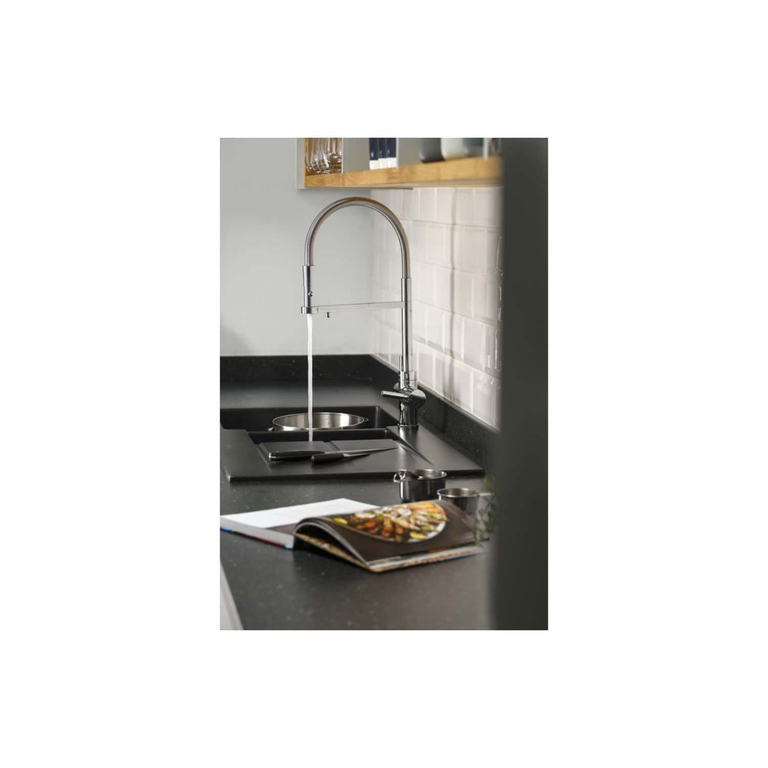 Modern kitchen sink with chrome faucet and black countertop, open cookbook and glassware on wooden shelf, white tiled wall, stylish and functional kitchen design.