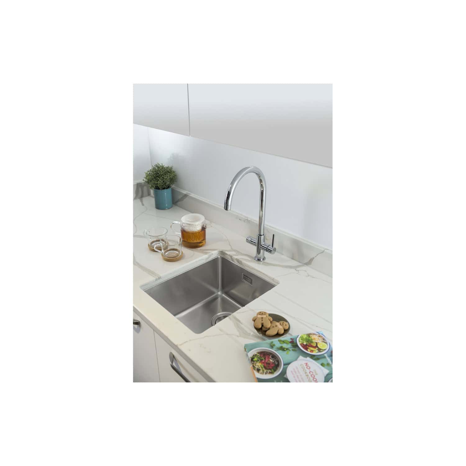 Wideshot of a modern kitchen sink with a sleek chrome faucet, set in a white marble countertop, with tea and biscuits, a potted plant, and a minimalistic backsplash, showcasing elegant bathroom and kitchen design.