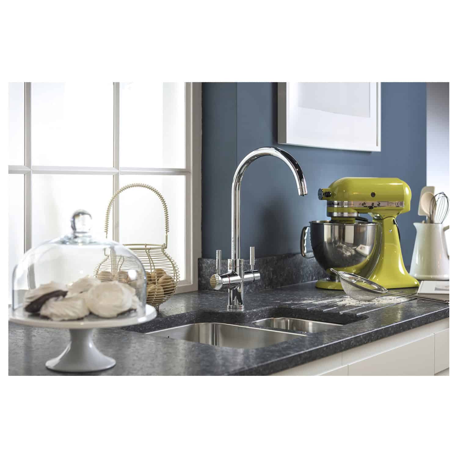 Sleek modern kitchen sink with chrome tap, surrounded by kitchen appliances including a lime green stand mixer and accessories like a wire basket and cake stand, in a stylish kitchen setting.