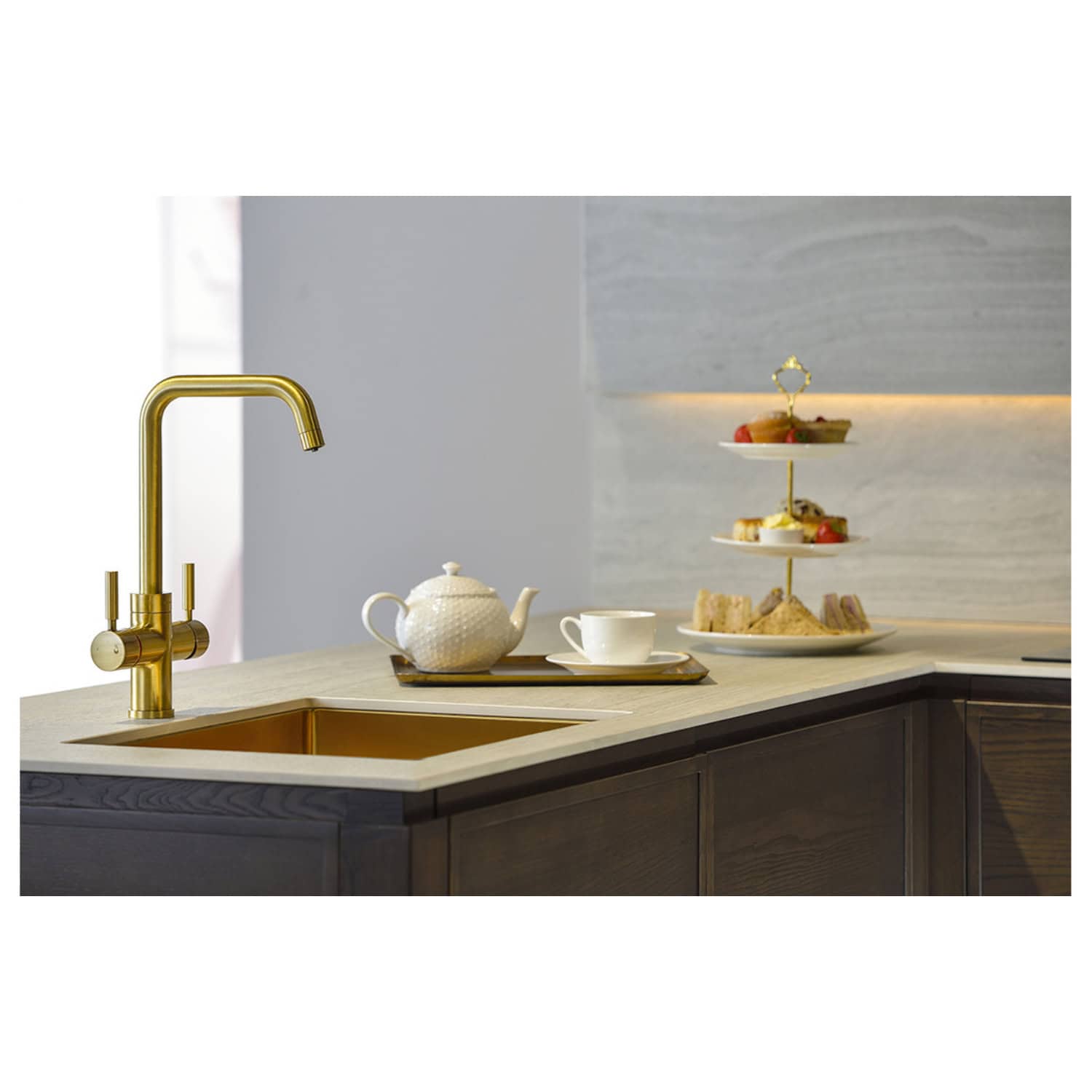 Elegant modern kitchen with a gold sink faucet, white tea set, and a three-tiered dessert tray on a beige kitchen island, showcasing luxury kitchen appliances and stylish decor.