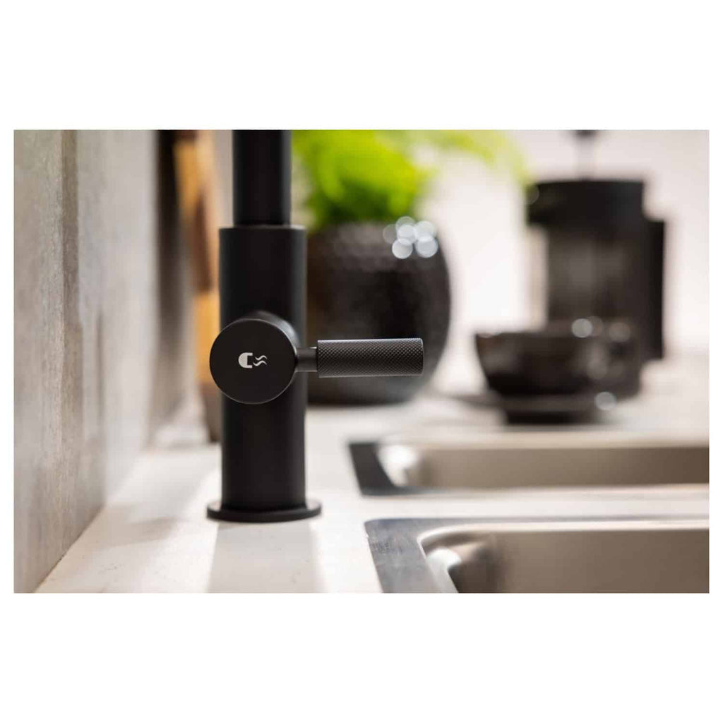 Sleek black kitchen tap with a modern design, featuring a textured handle and a minimalist logo, installed on a white countertop with a blurred background of a potted plant and kitchen essentials.