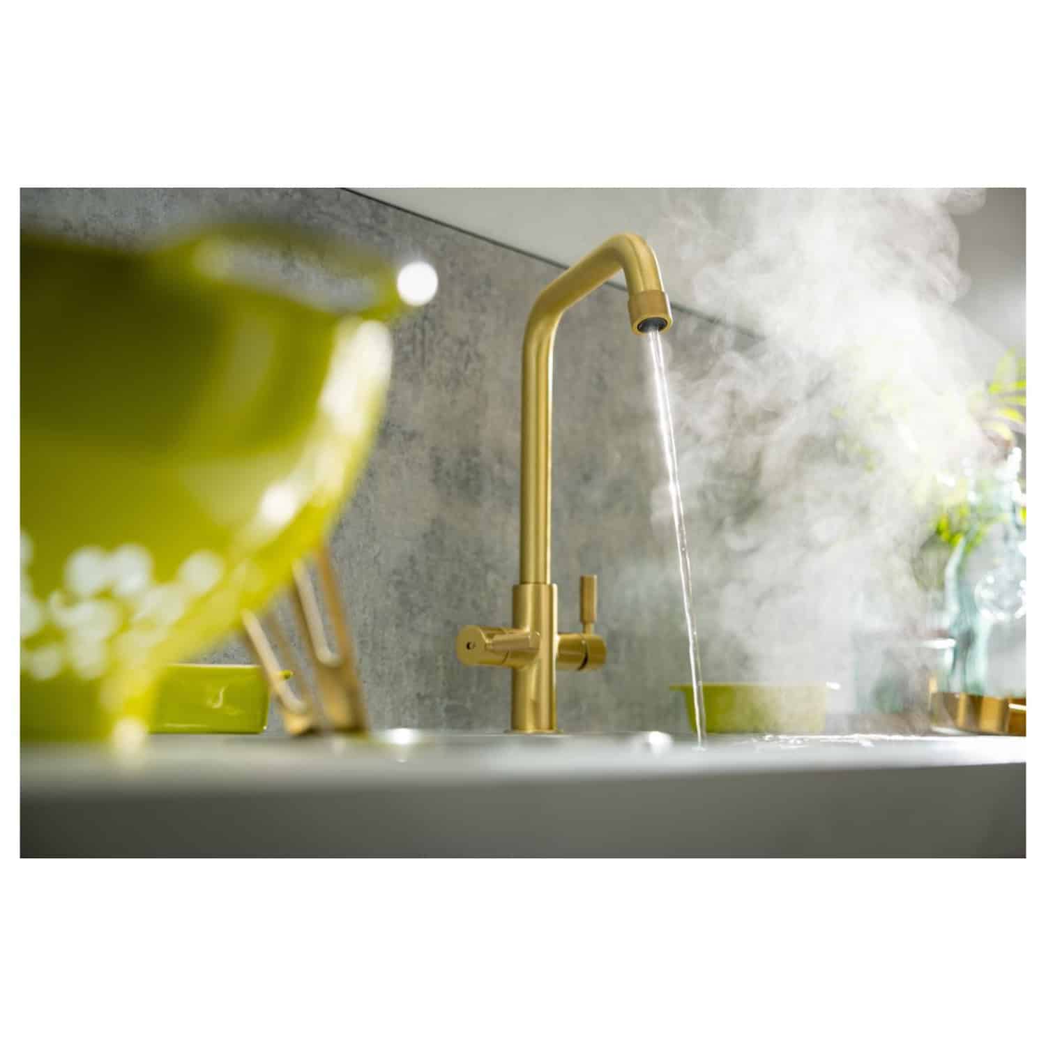 Shiny gold modern kitchen faucet with running water, stylish kitchen sink with green and yellow dishware, steam rising, sleek grey backsplash, perfect for contemporary bathroom or kitchen renovation.